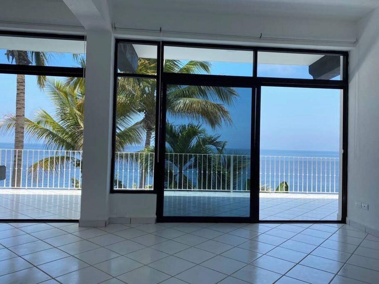 A room with large windows, white tiled floor, and a view of the sea and palm trees.