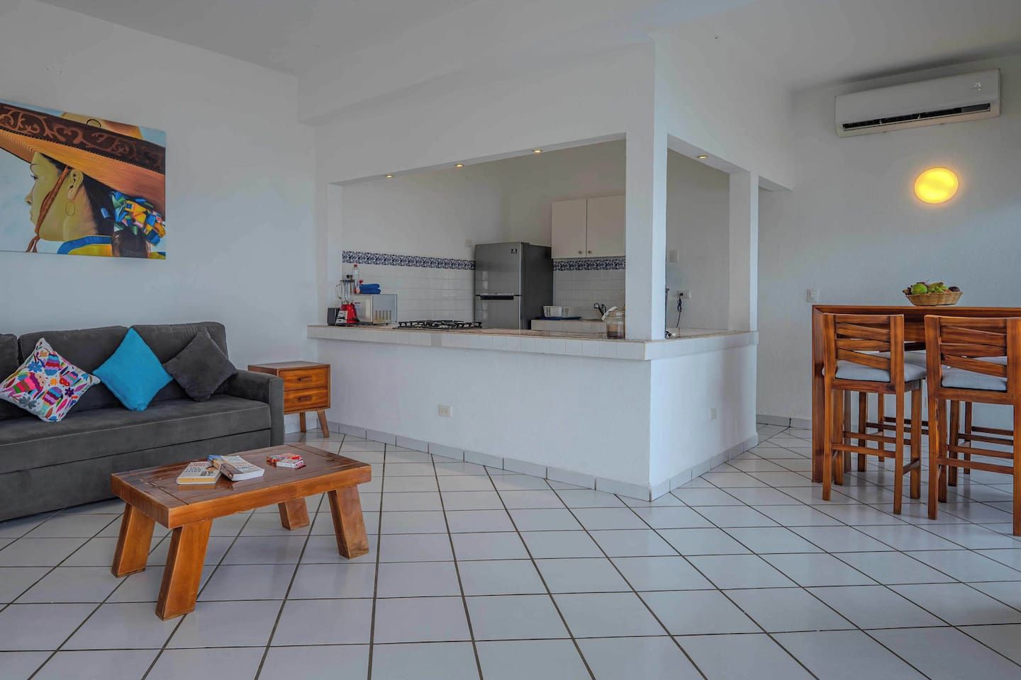 Uma sala de estar moderna com sofá cinza, mesa de centro de madeira e área de cozinha com armários brancos e painel de azulejos.