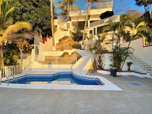 Une piscine dont le fond est recouvert de carreaux formant un motif géométrique, entourée d’une terrasse en béton, de palmiers et d’un immeuble à plusieurs étages doté de balcons.