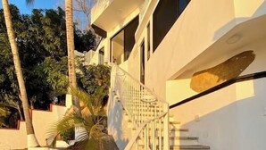 A white building with a staircase leading to a balcony. There are palm trees and a rock formation nearby. - Condo 1 at Del Mar PV (Aguacate)