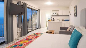 A hotel room with a bed, a gray sofa, a kitchen area, and a balcony with curtains. - Condo 1 at Del Mar PV (Aguacate)