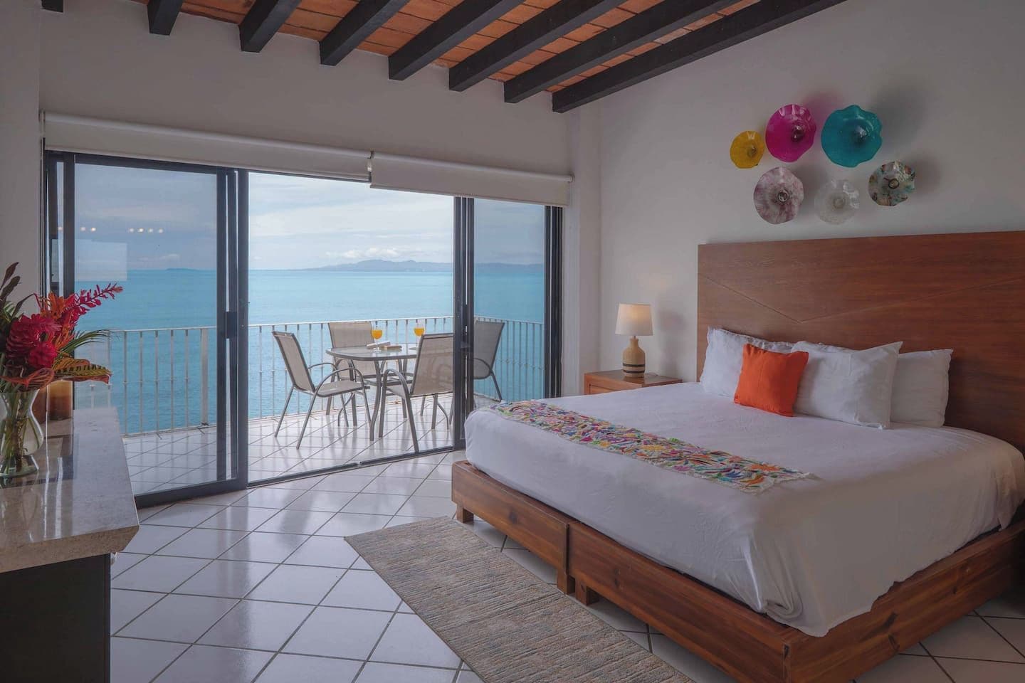 A bedroom with a large bed, a balcony with a table and chairs, and a view of the sea.