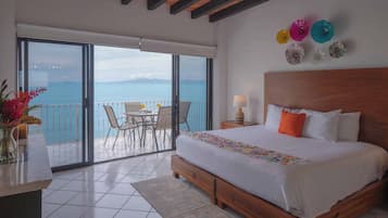 A bedroom with a large bed, a balcony with a table and chairs, and a view of the sea.