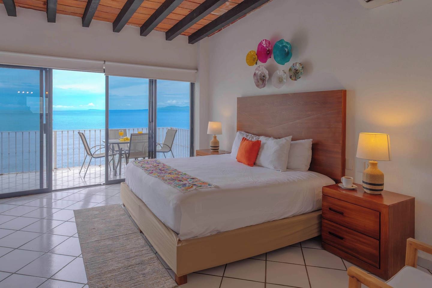 A hotel room with a large bed, a wooden headboard, a nightstand with a lamp, a small table with a cup, and a balcony with a table and chairs.