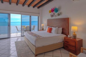 A hotel room with a large bed, a wooden headboard, a nightstand with a lamp, a small table with a cup, and a balcony with a table and chairs. - Del Mar PV Condo 2: Oceanfront 2BR, Pool & Slide (Aguacate)