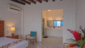 A hotel room with a bed, a chair, a nightstand with a lamp, an air conditioner, a bathroom with a sink and mirror, and a view of the sea. - Oceanfront Luxury Condo w/ Pool & Balcony Views (Aguacate)
