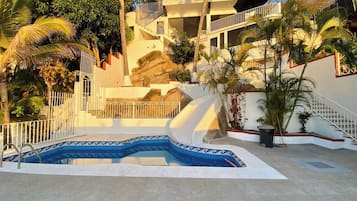 A swimming pool with a geometric tile pattern, surrounded by a concrete deck, palm trees, and a multi-story building with balconies.