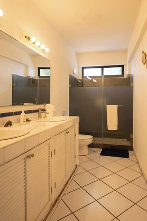 A bathroom with a white sink, mirror, and toilet, a shower area, and a tiled floor. - Del Mar PV Condo 2: Oceanfront 2BR, Pool & Slide (Aguacate)