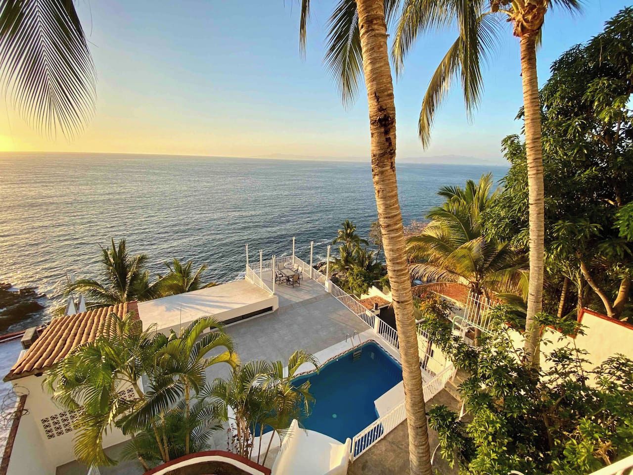 A pool area with a view of the sea, surrounded by palm trees and a white building.