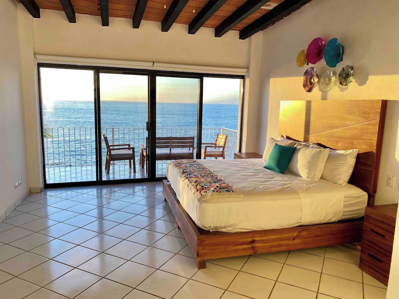 A bedroom with a large bed, a view of the sea through a sliding door, and a balcony with chairs.