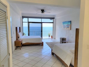 A bedroom with two beds, a ceiling fan, a large window with a view of the sea, and a small table with a chair. - Oceanfront Villa: Private Resort w/ Heated Pools (Aguacate)