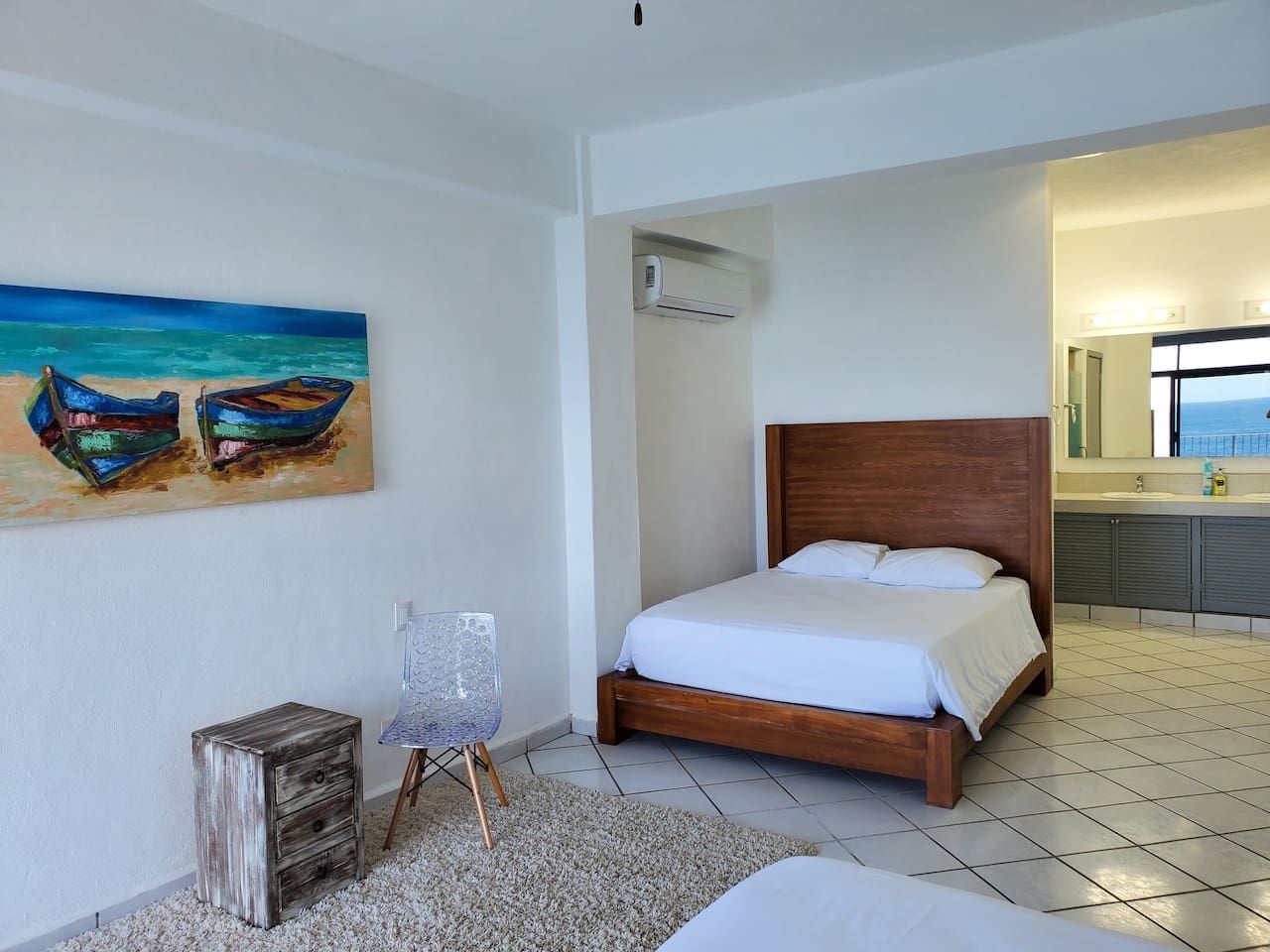 Quarto com cama, pintura de barcos, uma cadeira e um pequeno armário de madeira.