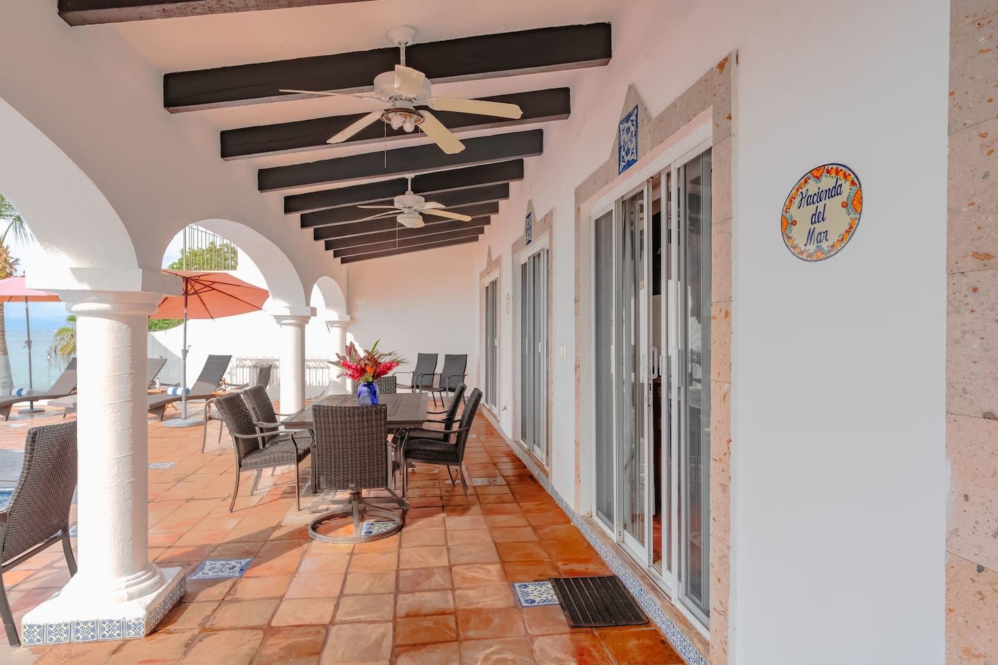 A covered outdoor seating area with tables and chairs, ceiling fans, and a view of the sea.