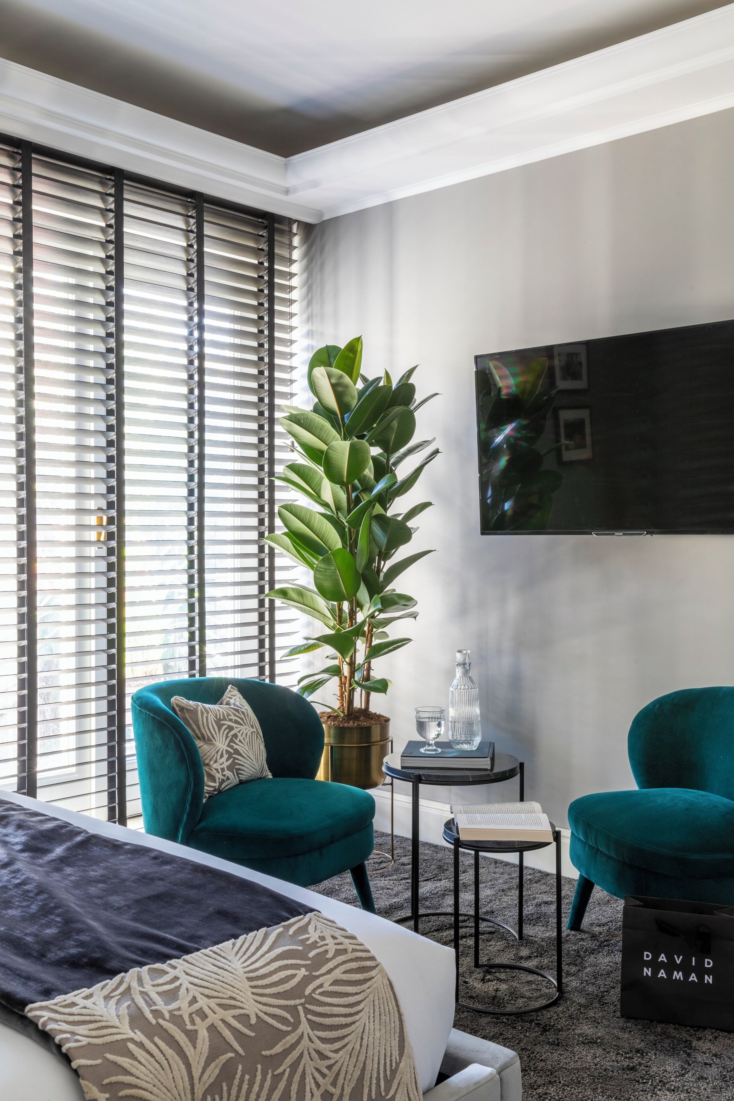 Moderni hotellihuone, jossa on suuri sänky, turkoosinväriset nojatuolit, ruukkukasvi, pieni pöytä, jonka päällä on kirja ja lasi, sekä seinälle asennettu taulutelevisio.
