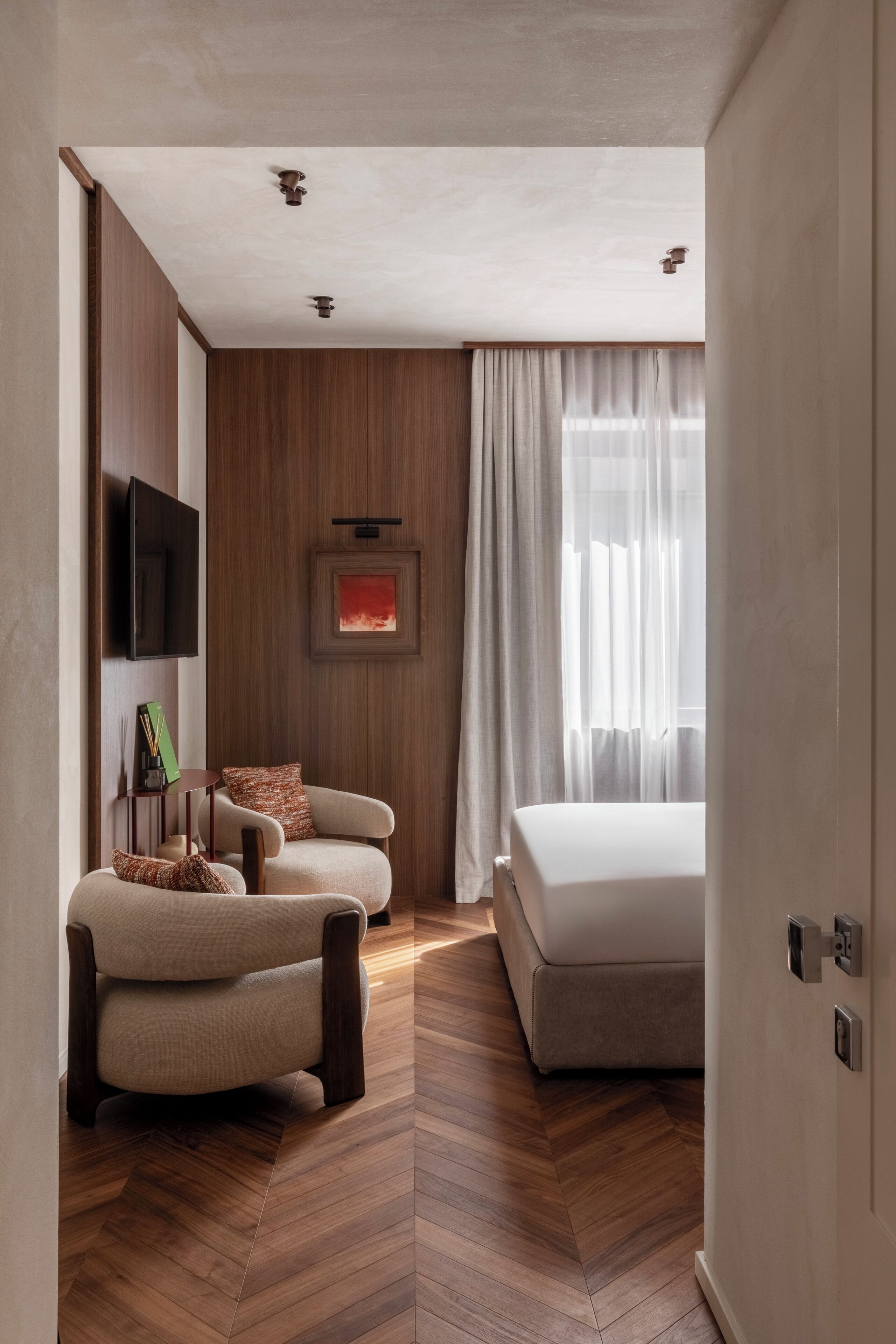 Une chambre d’hôtel moderne avec un plancher en bois, un canapé, un lit, un téléviseur fixé au mur et une fenêtre avec des rideaux.