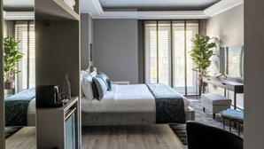 A modern bedroom with a large bed, bedside tables, a desk, and a chair. There are two potted plants and a large window with blinds. - Naman Hotellerie - Spagna (Rome)