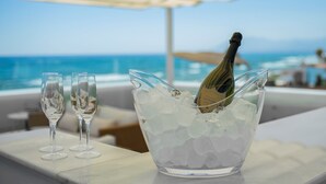 Una botella de champán en un cuenco de vidrio lleno de cubitos de hielo, con dos copas vacías a un lado, sobre una mesa con vistas al océano.