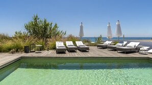 Una piscina cristalina con sillas de descanso y sombrillas, una terraza de madera y vista al mar.