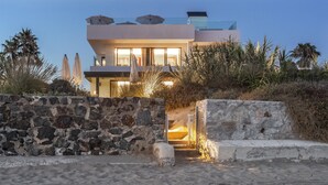 Una moderna propiedad frente a la playa con entrada de muro de piedra, zona de piscina y un edificio con ventanas amplias y sombrillas exteriores.