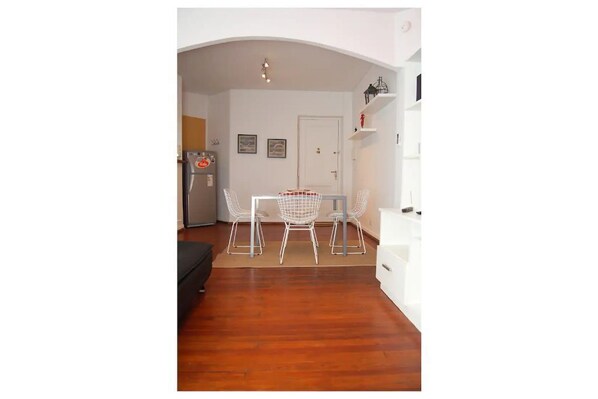 Uma área de refeições com mesa e cadeiras, um refrigerador e uma porta branca ao fundo.