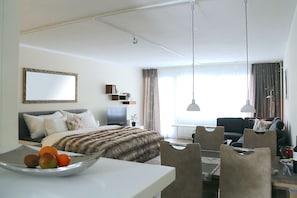 A modern hotel room with a bed, a desk with a bowl of fruit, a television, and a seating area with a sofa and chairs.