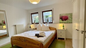 Une chambre à coucher avec un lit en bois, un tapis vert, une table de chevet avec une lampe, un miroir et une fenêtre donnant sur un paysage verdoyant.
