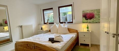 A bedroom with a wooden bed, a green rug, a bedside table with a lamp, a mirror, and a window with a view of greenery.