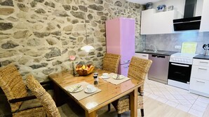 Un coin repas avec un mur en pierre, une table en bois, des chaises en osier, un réfrigérateur rose et une cuisine en arrière-plan.
