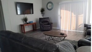 A living room with a grey sofa, a coffee table, a TV, a clock, and a sliding glass door. - Peoria Corner Lot 3 Bedroom 2 Bath with High Speed Internet/Smart TVs (Peoria)
