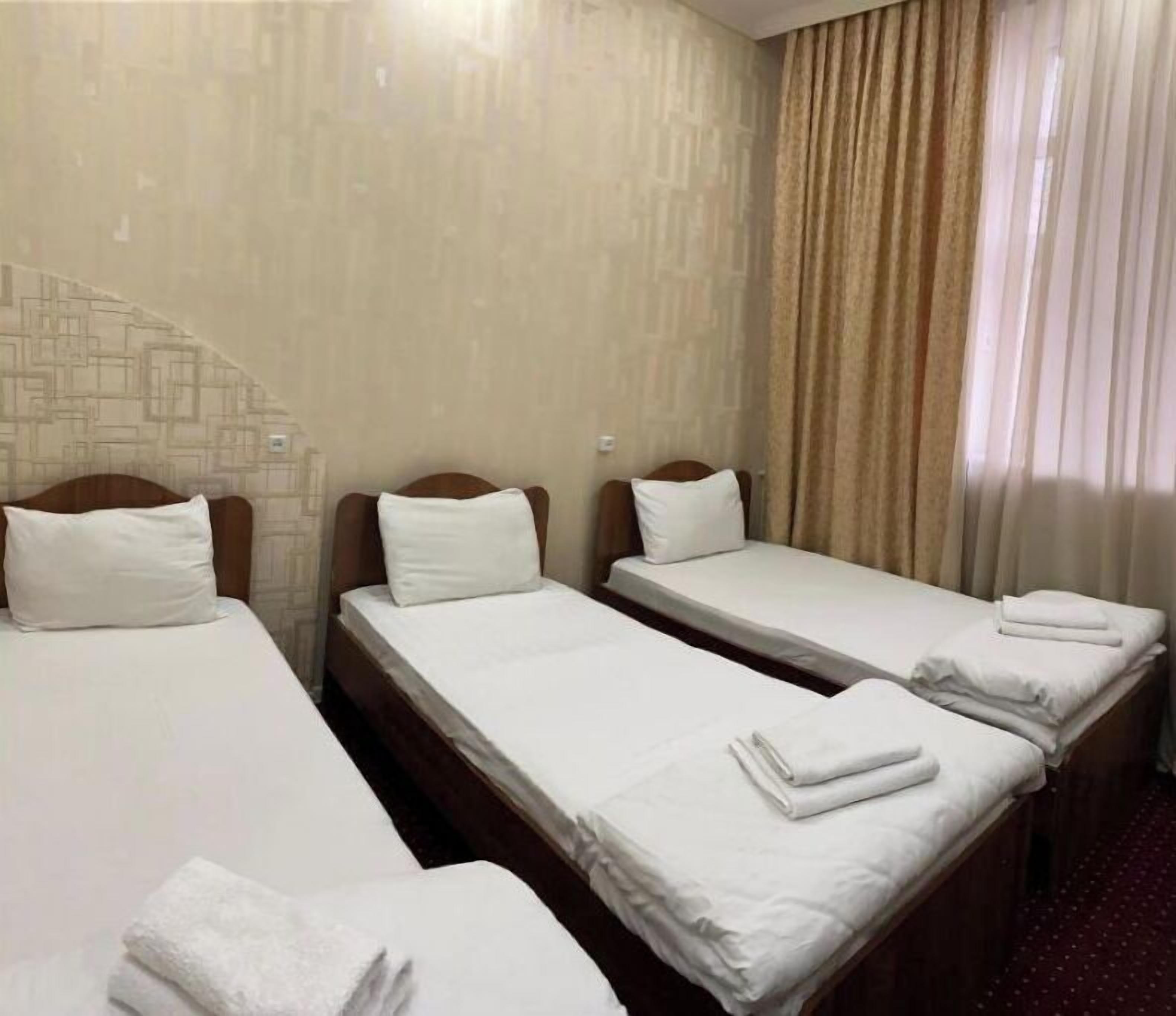 Habitación de hotel con tres camas individuales, cada una con sábanas y almohadas blancas. La habitación tiene una pared con diseño y una ventana con cortinas.