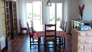 Un comedor con una mesa dispuesta para cuatro personas, rodeada de sillas. A la derecha, un pilar de piedra y, encima de la mesa, un estante de madera. A la izquierda, una puerta con un diseño de celosía.