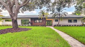 A modern house with a large tree in the front yard, a paved walkway, and a well-maintained lawn. - Retro St Pete Home w/ Private Heated Pool (St. Petersburg)