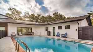 A swimming pool with a tiled deck, a covered patio area with chairs, and a fenced backyard. - Retro St Pete Home w/ Private Heated Pool (St. Petersburg)