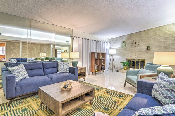 Ein Wohnzimmer mit blauem Sofa, einem Holztisch und einem gemusterten Teppich. An der Wand befinden sich ein Bücherregal und ein Spiegel.