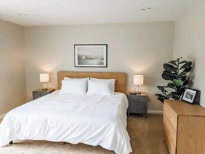 Cama bem arrumada com lençóis brancos, duas mesas de cabeceira com luminárias, um quadro emoldurado na parede e uma grande planta verde sobre um aparador de madeira.