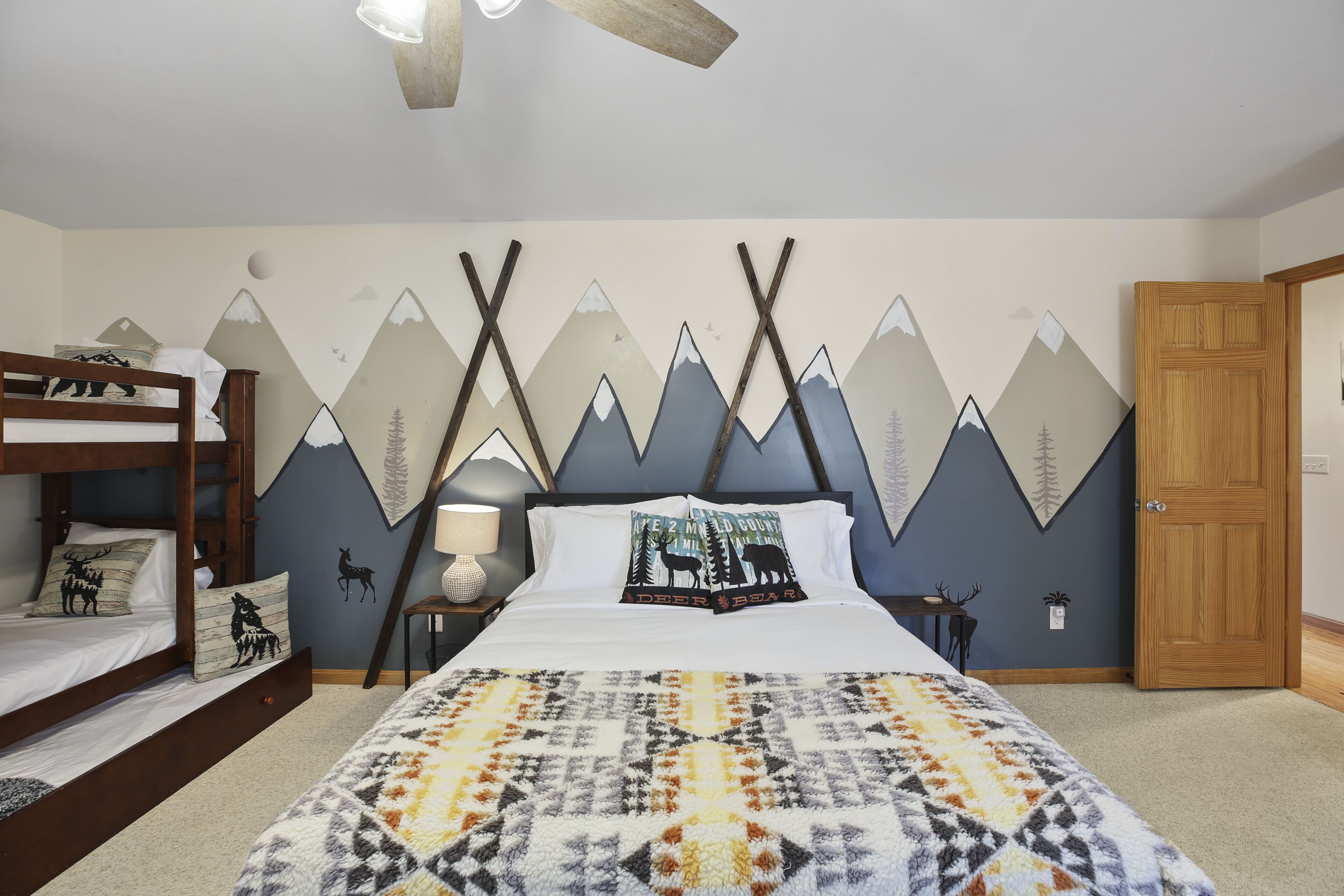 A bedroom with a bunk bed, a bed with mountain-themed wall art, a patterned blanket, and a wooden door.
