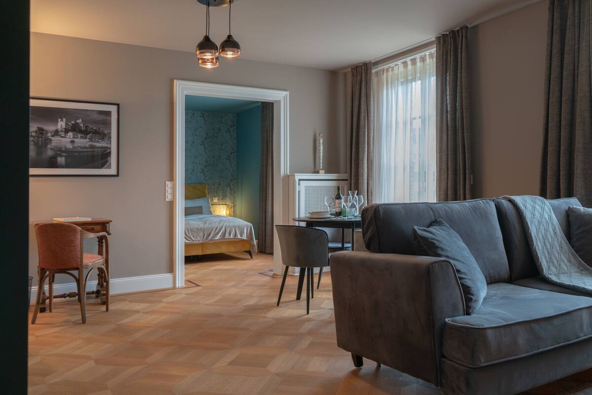 A modern living room with a grey sofa, a wooden floor, a bed, a desk, and a chair.
