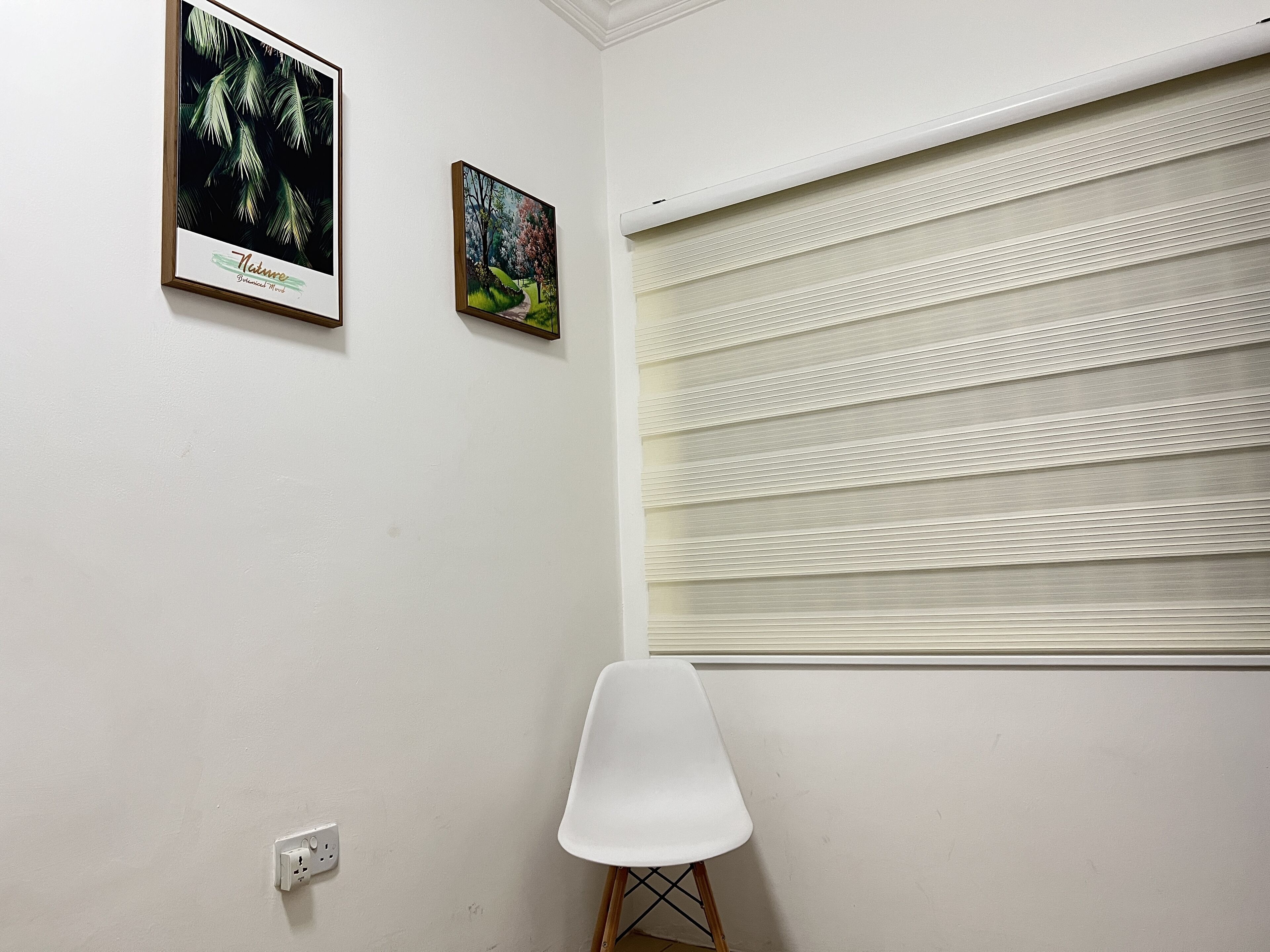 Una habitación con pared blanca, dos cuadros enmarcados, una silla blanca y una ventana con persianas horizontales.