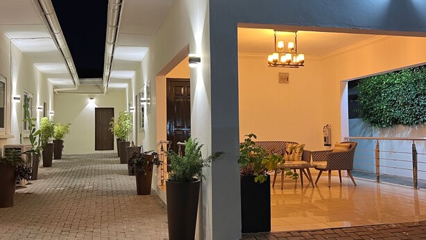 A well-lit outdoor area with a brick pathway leading to a seating area with a table and chairs. There are potted plants along the pathway and a wall with a green plant on the right. - Quiet escape, good security and serene environment (Accra)
