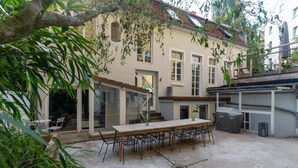 Un'area patio con tavolo e sedie in legno, una griglia coperta e un edificio con ampie finestre e un balcone.