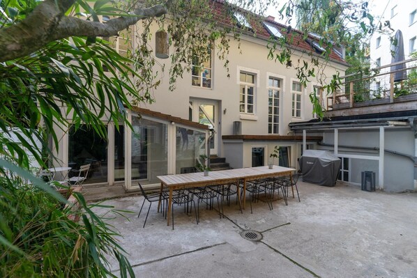 Un patio con mesa y sillas de madera, una parrilla cubierta y un edificio con ventanas amplias y un balcón.
