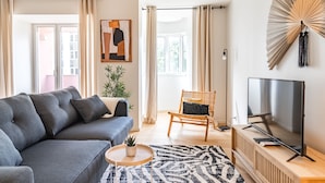 A modern living room with a grey sofa, a wooden chair, a TV on a wooden stand, and a large window with curtains.
