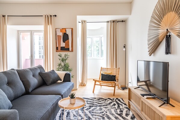 Un salon moderne avec un canapé gris, une chaise en bois, un téléviseur posé sur un meuble en bois et une grande fenêtre avec des rideaux.