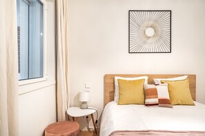 Une chambre d’hôtel bien rangée, avec un lit, une table de chevet dotée d’une lampe, un petit tabouret rond, une fenêtre avec des rideaux et une œuvre d’art murale.