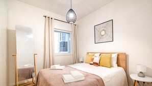 A neatly arranged hotel room with a bed, a bedside table, a mirror, a window with curtains, and a wall art piece.