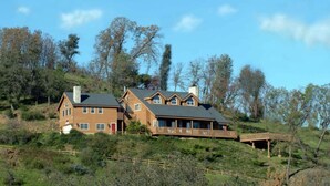 Une grande maison en bois à deux étages, dotée d’un porche couvert et d’un petit pont qui la relie à un deuxième bâtiment.