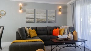 Een moderne woonkamer met een grijze bank, een bijzettafeltje en decoratieve objecten. Er hangen wandlampen en kunstwerken boven de bank.