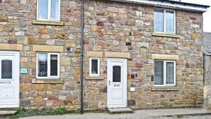 A stone building with three white-framed windows and doors, a visible number plate, and a small overhang above the windows. - Braidcarr Cottage (Seahouses)