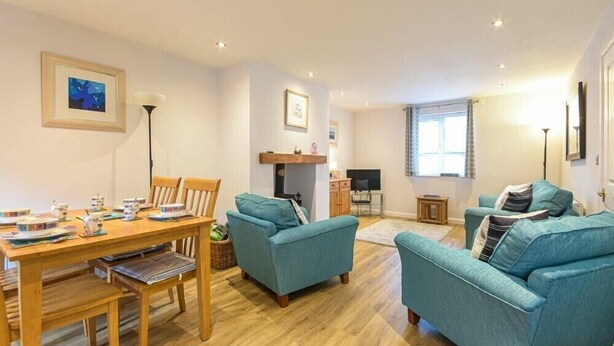 A living room with a wooden dining table, teal sofas, and a fireplace. - Braidcarr Cottage (Seahouses)