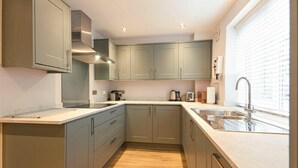 A compact kitchen with grey cabinets, a stainless steel range hood, a built-in oven, and a window with blinds. - Braidcarr Cottage (Seahouses)